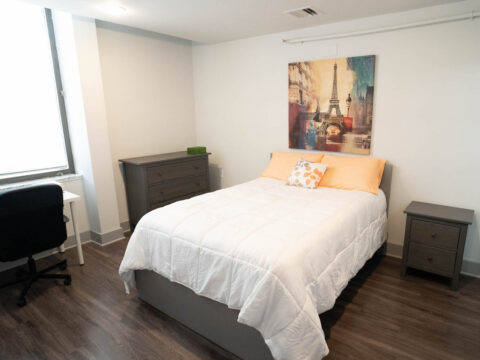 10th floor model bedroom showcasing nightstand, storage drawer, a desk in front of the windows and bed at the center of the wood floor room.